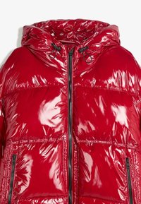 Giacca a vento rossa con finitura lucida, dotata di cappuccio, cordini regolabili e due tasche frontali con zip.