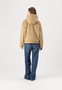 Beige hettegenser med en avslappet passform, laget av strukturert materiale, kombinert med mørkeblå vide jeans. Jakken har croppet lengde.