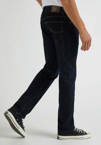 Jeans in denim scuro con taglio dritto, cuciture gialle, cinque tasche e un'etichetta in pelle marrone sul retro. Indossati con sneaker nere.