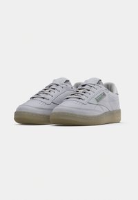 Light gray suede sneakers with a low-top design, perforated toe box, and gum rubber outsole. Features a green and red logo accent.