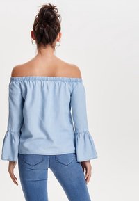 Lysblå off-the-shoulder top med lange, udvidede ærmer og elastisk halsudskæring. Båret med mørkeblå jeans, der viser en afslappet stil.