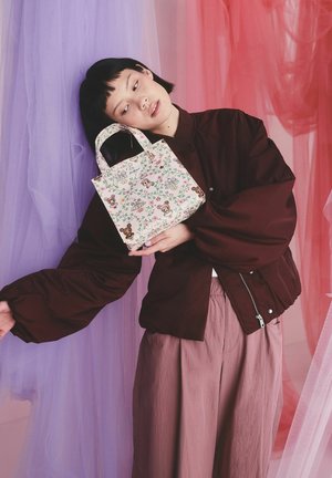 Mladá osoba v oversize tmavej bunde a fialových nohaviciach drží kvetinovú tašku s kreslenými medvedíkmi na pozadí z ružovej a fialovej priehľadnej látky.