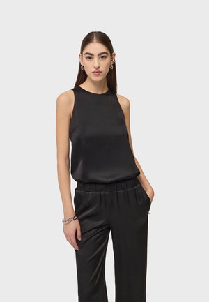 Kobieta o długich ciemnych włosach ubrana w czarny satynowy bezrękawnik i pasujące spodnie, stoi z jedną dłonią w kieszeni na jednolitym tle.