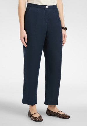 Donna che indossa pantaloni su misura blu scuro con tasche laterali, una maglia bianca, scarpe piatte intrecciate marroni e un braccialetto marrone a un polso.