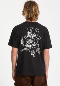 Person with shoulder-length blond hair wearing a black t-shirt featuring a white skull in a top hat smoking cigars with rolling dice design.