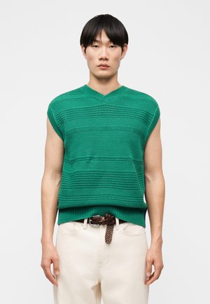 Jeune homme portant un pull sans manches vert en tricot avec une texture à motifs, associé à un pantalon blanc cassé et une ceinture tressée marron.