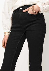 Jeans skinny en denim noir avec une taille mi-haute, une fermeture à bouton en métal argenté et un design traditionnel à cinq poches. Texture douce avec une légère élasticité.