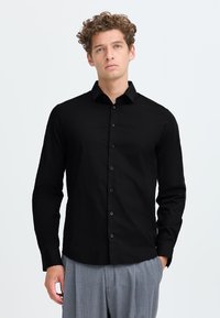 Giovane uomo con capelli ricci che indossa una camicia nera con bottoni e pantaloni grigi plissettati su uno sfondo bianco semplice.