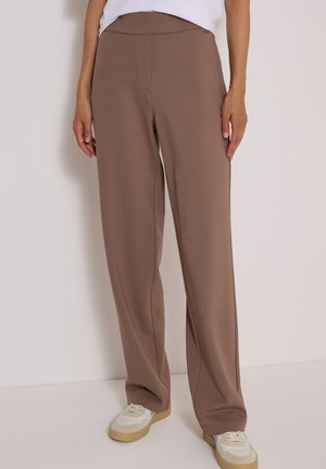 Personne portant un pantalon marron ample et des baskets blanches, debout devant un fond clair uni.