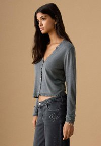 Cardigan gri scurtat cu decolteu ondulat, închidere cu nasturi și tiv zdrențuit, asortat cu blugi negri cu nituri decorative.