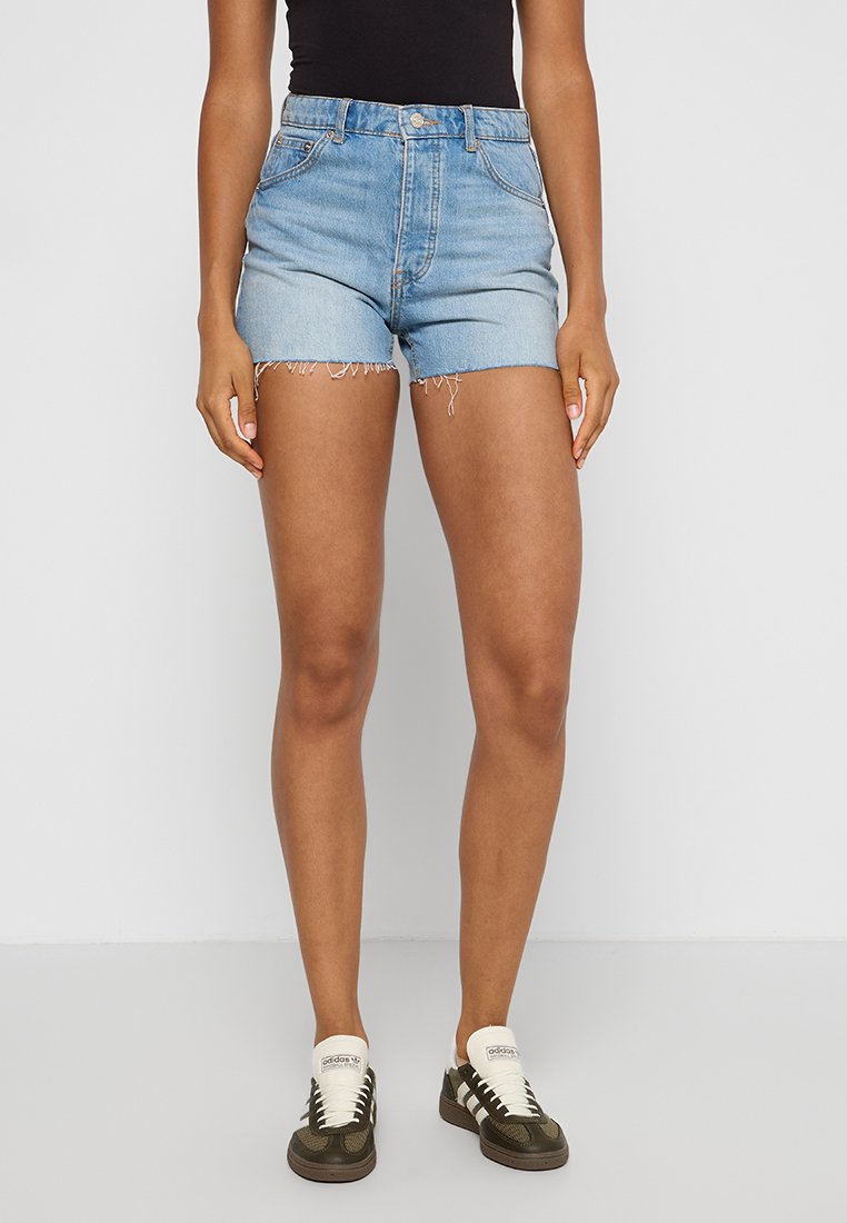 Shorts de mezclilla en azul claro con un dobladillo deshilachado y una cintura alta, combinados con zapatillas verdes y blancas, sobre un fondo liso.