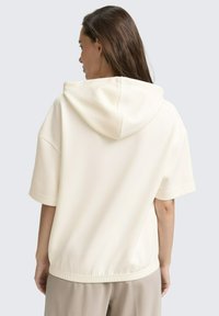 Cremefarbener Hoodie mit kurzen Ärmeln, Kapuze und elastischem Saum. Weiche Textur und minimales Design. Über hellbeigen Hosen getragen.