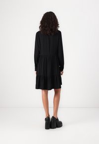 Abito lungo nero a maniche lunghe con vita arricciata e orlo a strati. Abbinato a scarpe con plateau nere. Tessuto liscio con un design semplice.