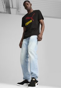 Fekete pamut póló piros Puma logóval és sárga szöveggel, világoskék farmernadrággal és fekete velúr sportcipővel, arany díszítéssel párosítva.