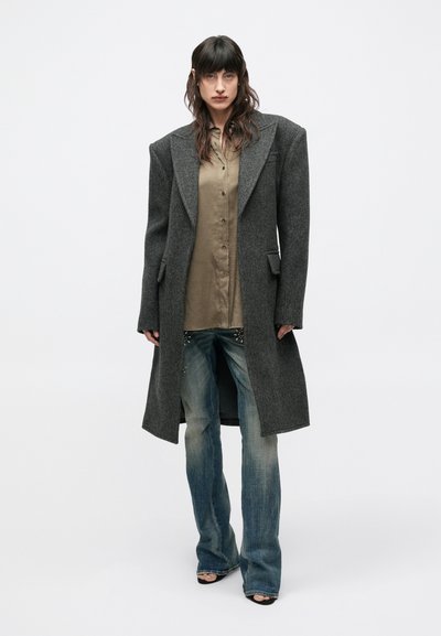 Grå, oversized uldfrakke med skarpe revers, kombineret med en beige skjorte med knapper og udvidede blå jeans med pynt nær kanten.