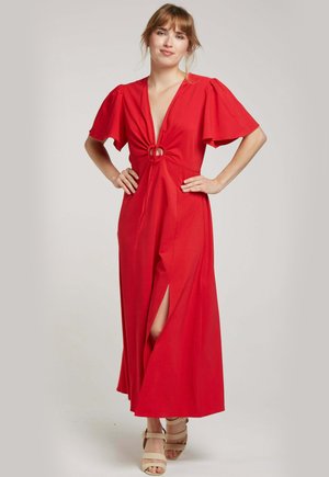Robe maxi rouge avec des manches courtes bouffantes, décolleté en V profond, détail de lien à la taille et fentes sur les côtés. Tissu lisse, coupe décontractée. Chaussures légères.