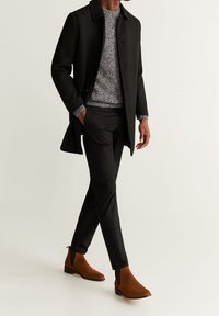 Cappotto di lana nero, maglione grigio a motivi, pantaloni neri su misura e stivali Chelsea in suede marrone, creando un look stratificato e sofisticato.