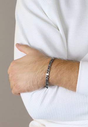 Silberfarbener Armreif mit Gliedern, die strukturierte Muster aufweisen. Er kontrastiert mit einem weißen, strukturierten Langarmshirt.