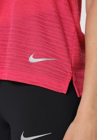 Różowa sportowa koszulka o fakturowanej powierzchni z zaokrąglonym dołem, z srebrnym logo Nike. Nosić z czarnymi legginsami, podkreślającymi dopasowany krój.