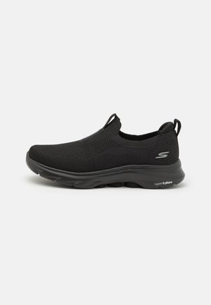 Črne slip-on športne copate s prozorno mrežasto zgornjimi deli, teksturirano zasnovo in oblazinjenim podplatom, ki ima zaobljen nos in subtilno blagovno znamko.