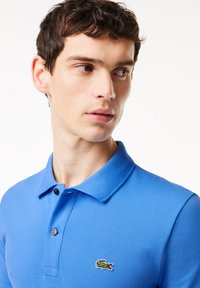 Blå polo-tröja i strukturerad bomull, med klassisk krage, två knappar och en grön krokodil-logotyp på vänster bröst.