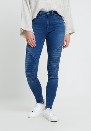Personne portant un jean skinny bleu style motard avec des panneaux côtelés aux genoux, des baskets blanches et un pull oversize gris clair.