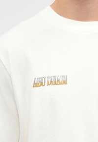 Λευκή αθλητική μπλούζα με κεντημένη την ένδειξη "ΑΜΠΟΥ ΝΤΑΜΠΙ" σε γκρι και πορτοκαλί. Κλασικό σχέδιο με λαιμόκοψη και μαλακή υφή, σε casual εφαρμογή.