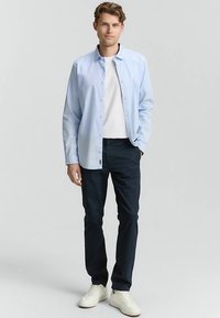 Svetlo modra srajca s sproščenim krojem in dolgimi rokavi, pod njo bela majica s krožnim izrezom, temne hlače in beli športni copati.