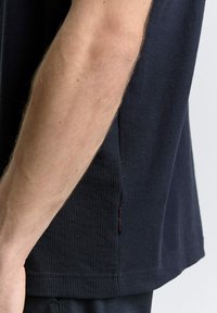 Navy tričko s vaflovou strukturou a krátkými rukávy, se bočním rozparkem a malou červenou štítkovou nášivkou na lemu. Minimalistický a neformální design.