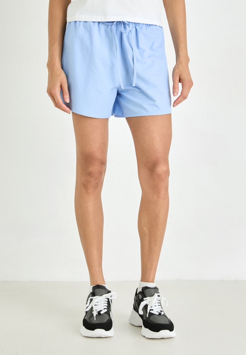 Pantaloni corto azzurri chiari con cintura elastica e laccio, caratterizzati da una texture liscia. Abbinati a sneakers nere e bianche.