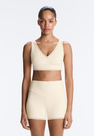 Kvinde iført beige sports-bh og matchende højtaljede biker-shorts, stående mod en ensfarvet lys baggrund, vendt fremad.