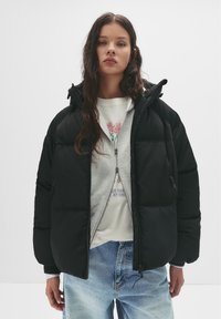 Crni zimski kaput s kapuljačom, s ukrašenim šavovima, zatvaranjem na patentni zatvarač sprijeda i elastičnim manžetama. Nositi preko svijetlosive dukserice s printom.