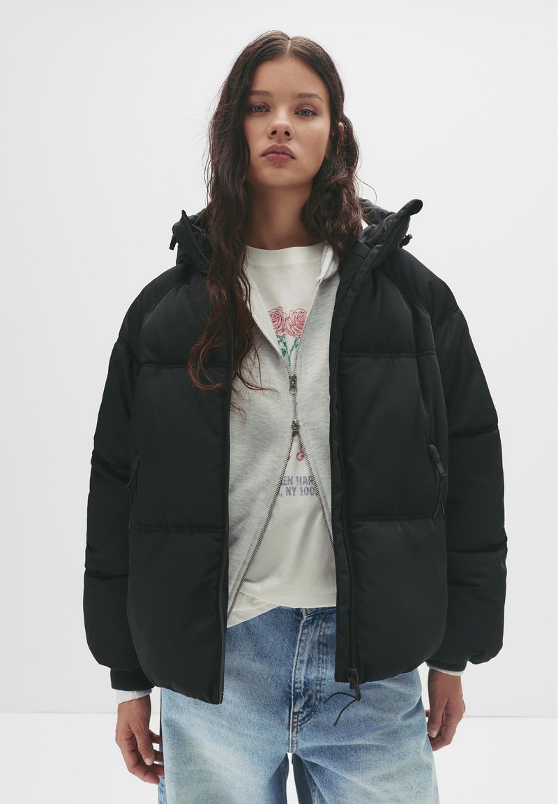 Crni zimski kaput s kapuljačom, s ukrašenim šavovima, zatvaranjem na patentni zatvarač sprijeda i elastičnim manžetama. Nositi preko svijetlosive dukserice s printom.