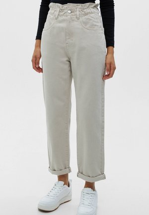 Kvinde iført beige højtaljede afslappede jeans rullet ved anklerne og hvide sneakers, sort langærmet top synlig ved taljen.