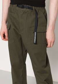Man in olijfgroene cargobroek met flapzakken en een zwarte riem met een label met de tekst "Converse".
