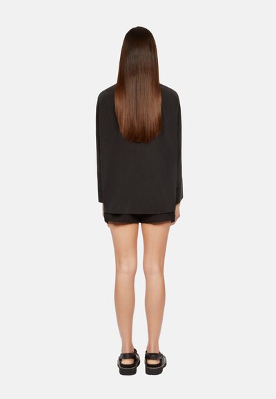 Zwarte oversized blazer met lange mouwen, gecombineerd met zwarte shorts. Gladde textuur en een strak ontwerp, bekeken vanuit de achterkant. Zwarte platform sandalen.