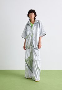 Veste argentée surdimensionnée avec un col pliable et un pantalon large assorti, tous deux en matériau réfléchissant et à coupe décontractée, sur un haut vert.