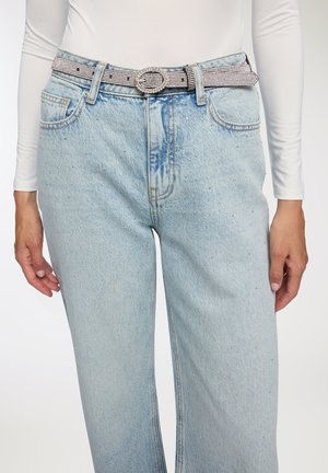 Persona che indossa jeans in denim azzurro chiaro con piccoli strass e una cintura argentata con strass sopra una maglietta bianca a maniche lunghe.
