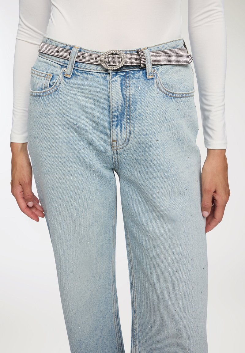 Persona che indossa jeans in denim azzurro chiaro con piccoli strass e una cintura argentata con strass sopra una maglietta bianca a maniche lunghe.