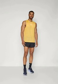 Κίτρινη αμάνικη μπλούζα με στρογγυλή λαιμόκοψη και λογότυπο, συνδυασμένη με μαύρα σορτς και ναυτικές αθλητικές παπούτσια. Απλός σχεδιασμός, αναπνεύσιμο υλικό.