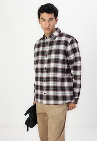 Camisa de franela a cuadros en blanco y negro con detalles rojos, diseño abotonado y un logo verde en el pecho izquierdo, combinada con pantalones beige.