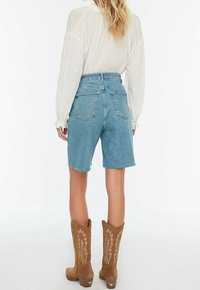 Mujer con shorts de mezclilla azul de talle alto, una blusa blanca de manga larga y botas vaqueras color canela con diseños de estrellas, de pie y de espaldas.