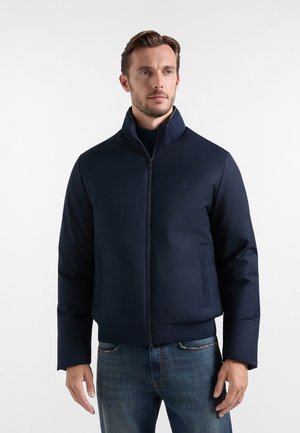 Uomo con giacca blu scuro con zip e collo alto e jeans blu, in piedi davanti a uno sfondo bianco semplice.