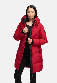 Woman wearing a long red padded winter coat with a hood, black zipper, and black clothing underneath, standing against a plain background.