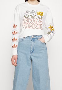 Vit långärmad skjorta med texten "ALWAYS ORIGINAL" och blommönster i gult och grönt, samt röda Adidas-logotyper på ärmarna. Jeanz i denim.