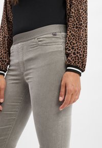 Graue Stretch-Jeans mit schlankem Schnitt und einem Bund, der den Markennamen trägt. Der Stoff hat eine glatte Textur, ergänzt durch ein Leopardenmuster-Hemd.