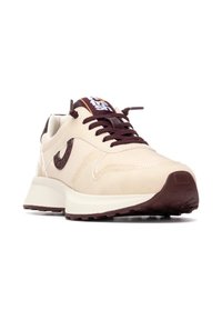 Zapatillas beige con suela acolchada en beige y marrón oscuro, tejido texturizado, cordones marrón oscuro y una etiqueta de la marca en naranja y blanco en la lengüeta.