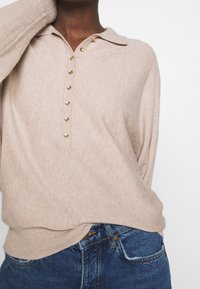 Maglione beige con colletto e patta con bottoni tono oro. Texture morbida e vestibilità comoda, abbinato a jeans denim blu.