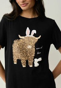 Černé tričko s grafikou huňatého hnědého skotského skotu s králičími ušima vedle malého bílého králíka. Na tričku je nápis "mooch love."
