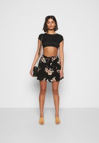 Top crop preto, saia com estampa floral e cintura com laço, e sapatos slip-on bejes. O conjunto apresenta um corte descontraído e tecido leve.